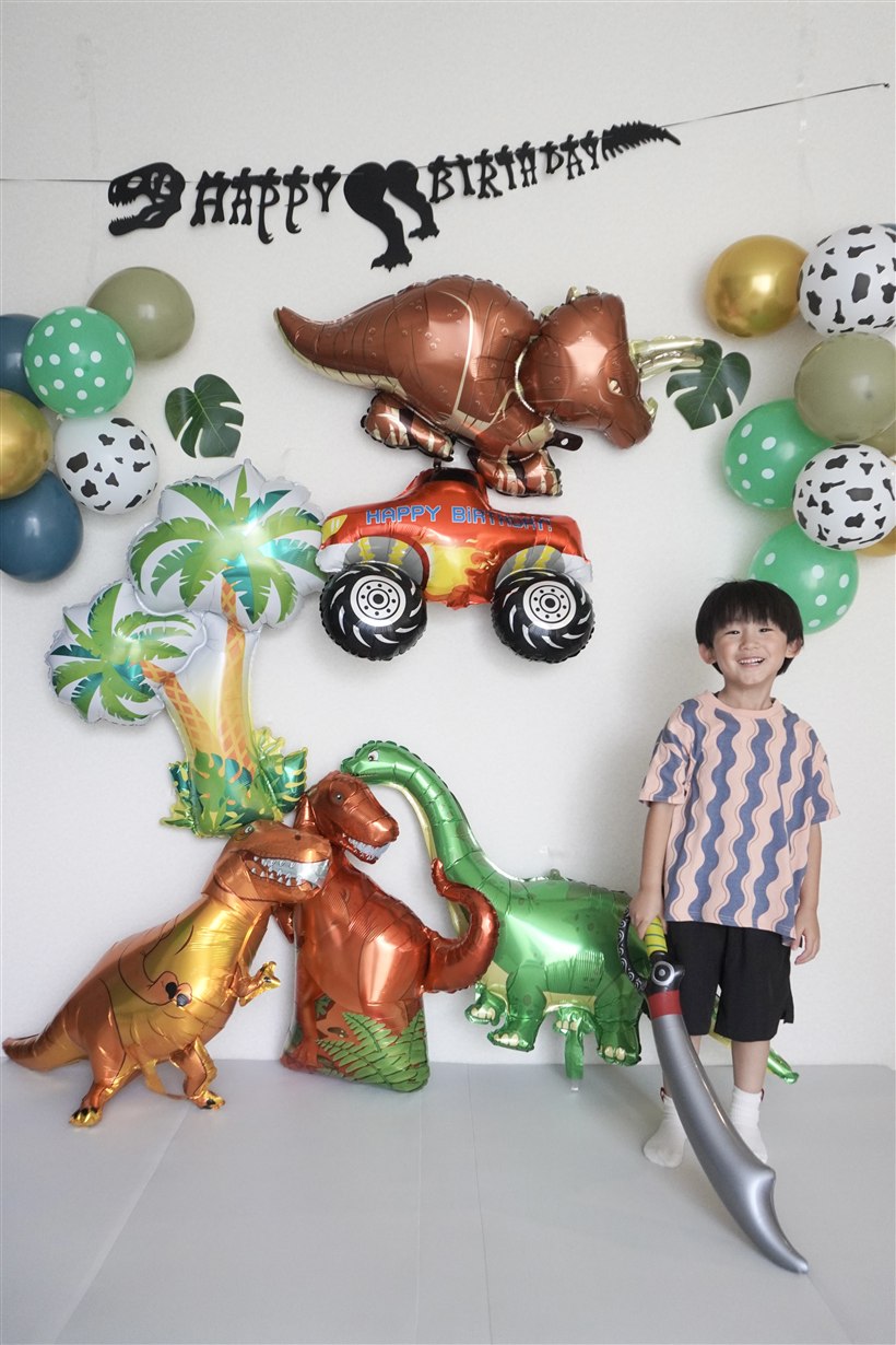 剣を手に立ち、笑顔を見せる4歳の男の子。恐竜バルーンとモンスタートラック、ヤシの木のバルーン