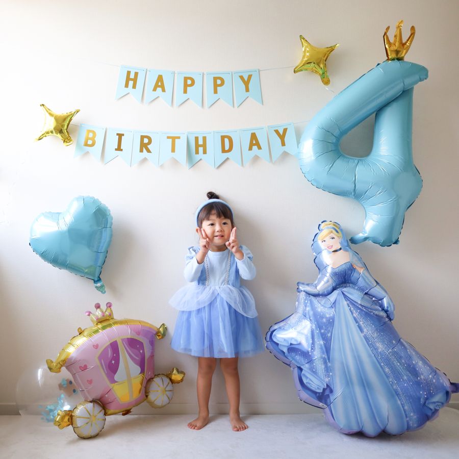 シンデレラのバルーンとかぼちゃの馬車のバルーンが床に置かれ、王冠つきの数字4と水色のHAPPY BIRTHDAYの紙のガーランド、ハート、星が壁の高い位置にある飾り付け。中央にドレスを着た子どもが立っている