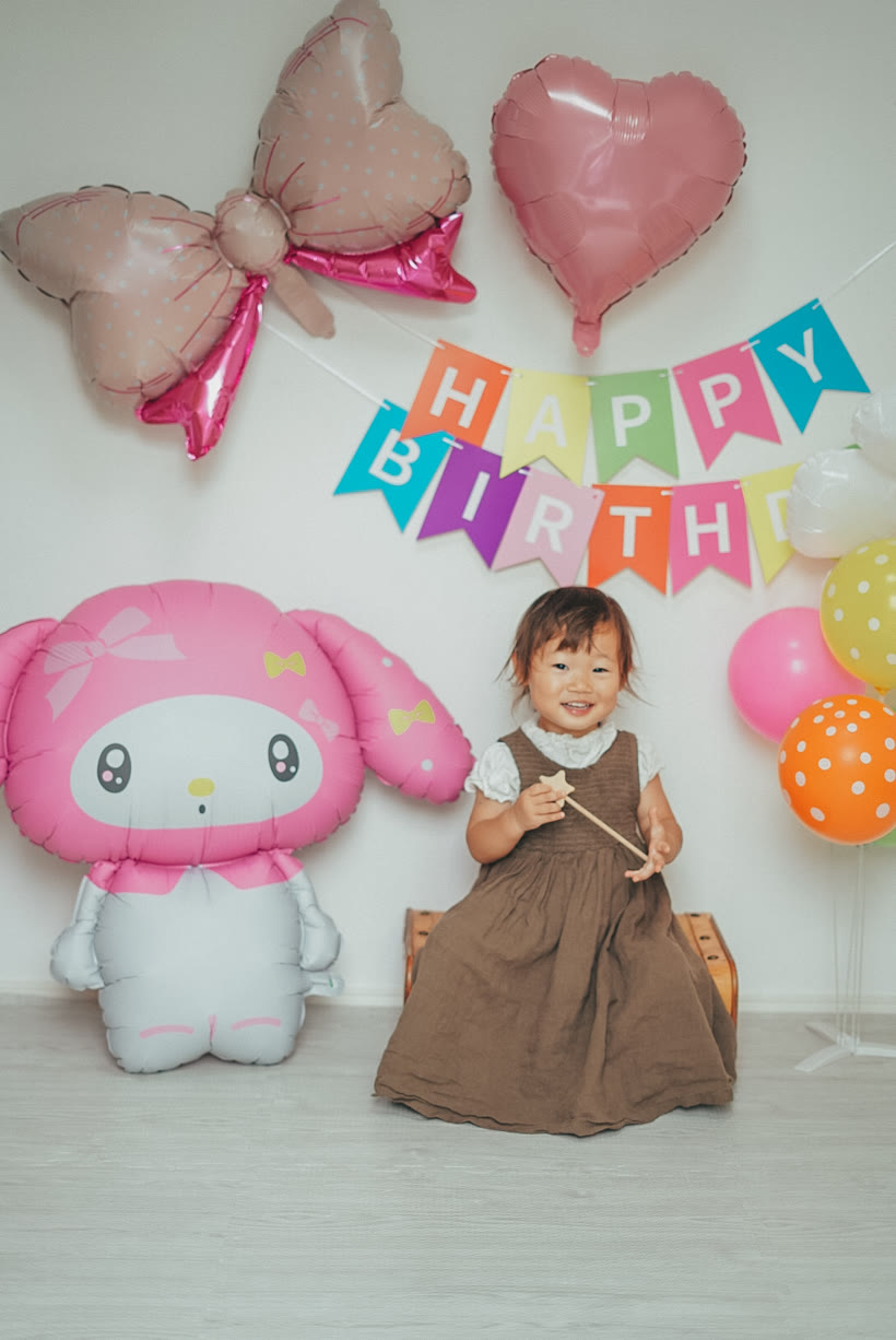 マイメロディのバルーンとカラフルなガーランドの前で、椅子に座って星のステッキを持つ2歳の女の子。やわらかいフィルム調の一枚