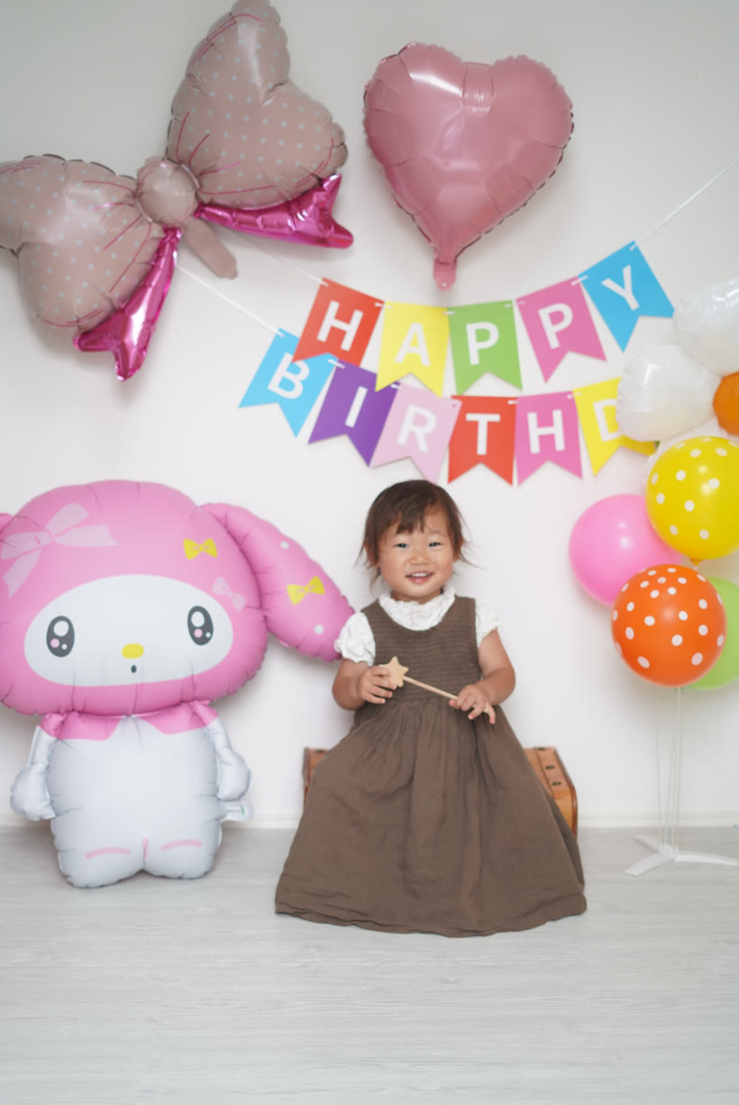 特大マイメロディバルーンの隣で、椅子に座ってにっこり笑う2歳の女の子。カラフルなHAPPY BIRTHDAYガーランドとリボン・ハートのバルーン、バルーンスタンドに浮かせた水玉のゴム風船