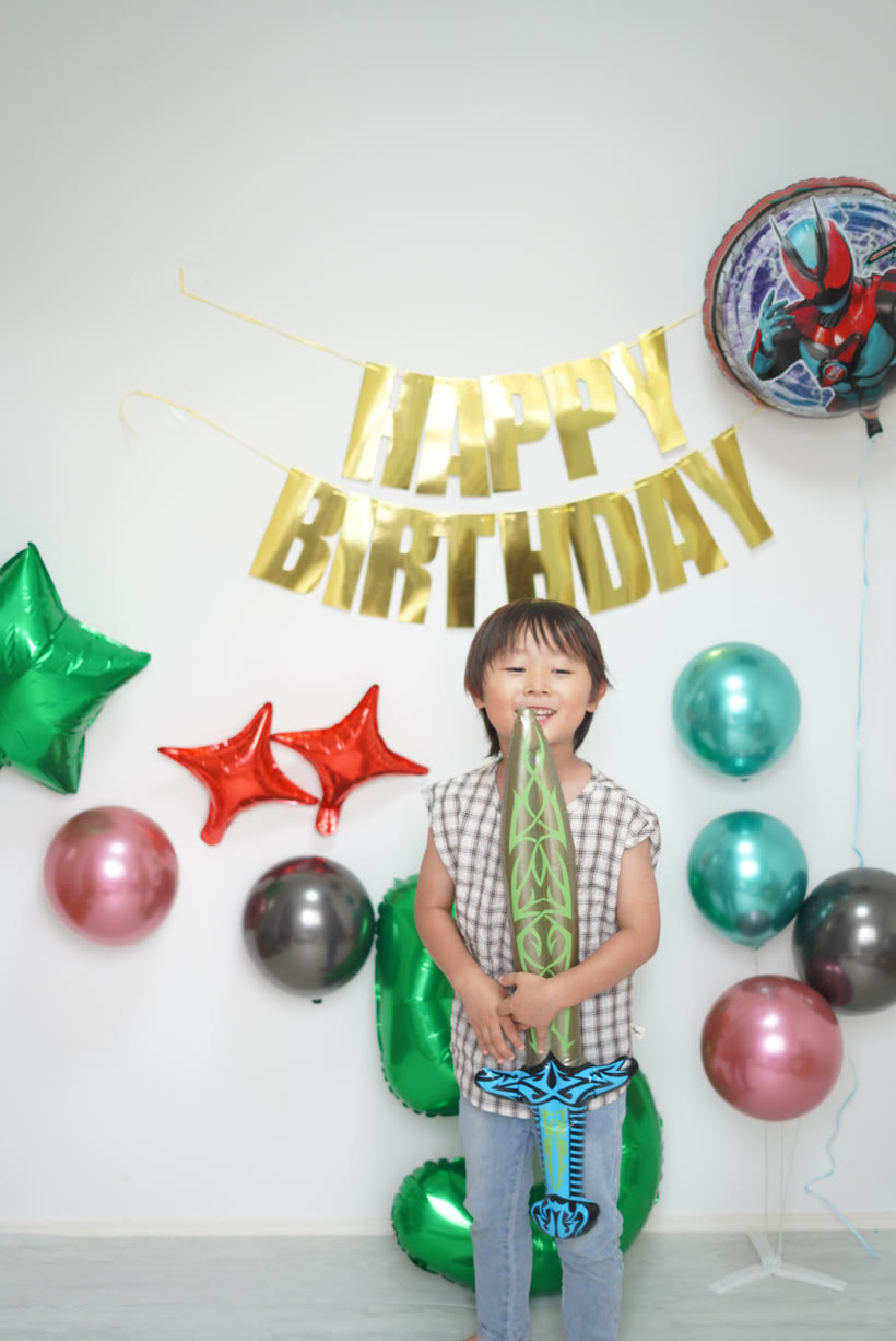 仮面ライダーゼッツのバルーンと金のHAPPY BIRTHDAYガーランド、緑の数字バルーン5の前で、付属の剣を構えて笑う5歳の男の子