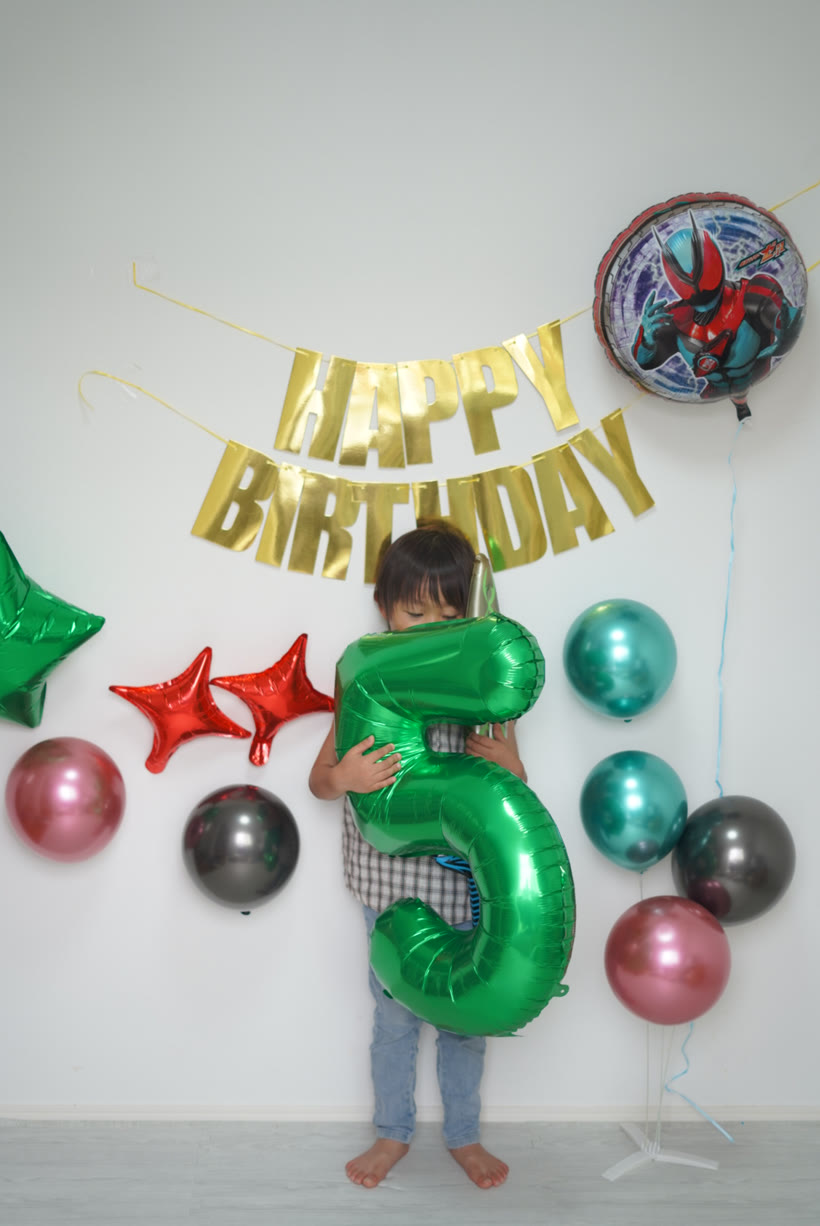 自分の身長ほどの大きな緑の数字バルーン5をぎゅっと抱きしめる5歳の男の子