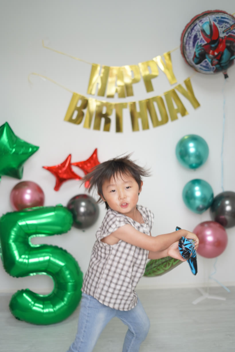 髪をなびかせて剣のバルーンを本気で振り抜く5歳の男の子。まわりにメタリックのゴム風船と緑の数字バルーン5