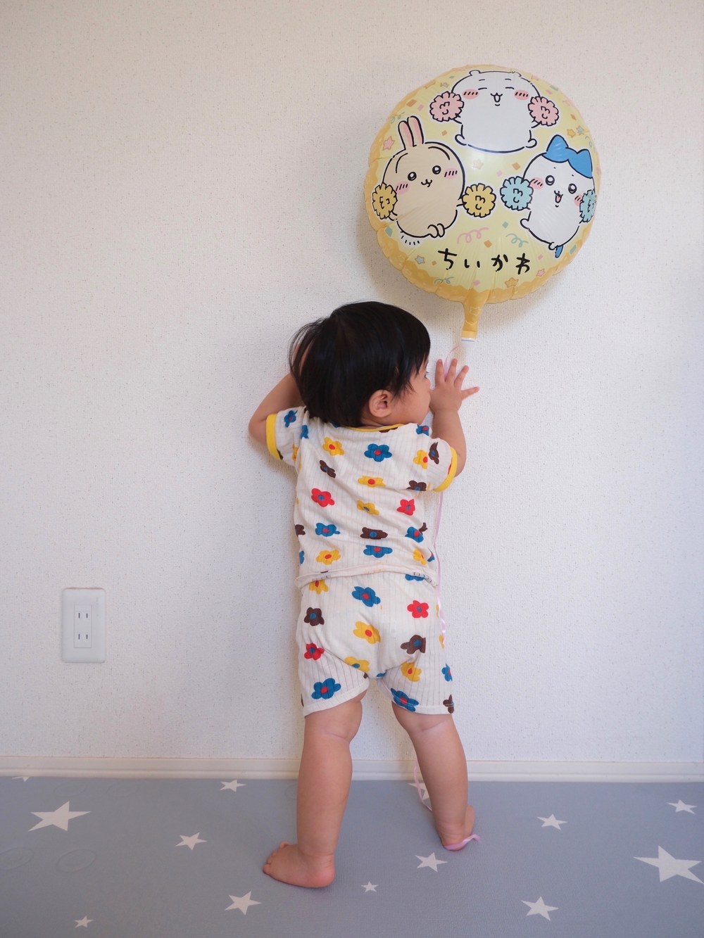 白い壁に貼ったちいかわ・ハチワレ・うさぎの丸いバルーンの下で、後ろ姿のお子さまが壁に両手をつき、右手をバルーンの根元へのばしている。花柄のお洋服、床は星柄のマット