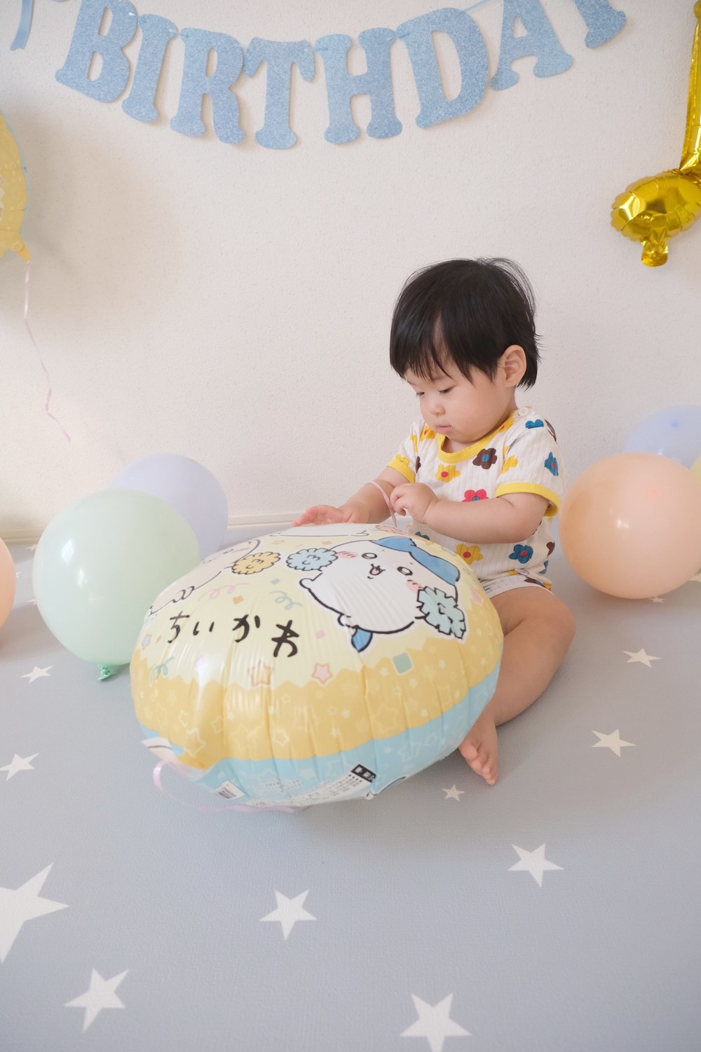 床に座ったお子さまが、ちいかわと書かれた黄色い丸いバルーンに手をのせて、リボンをつまみながら、じっと見ている。白地に赤・黄・青の花柄、襟と袖が黄色のお洋服。まわりにパステルカラーの風船
