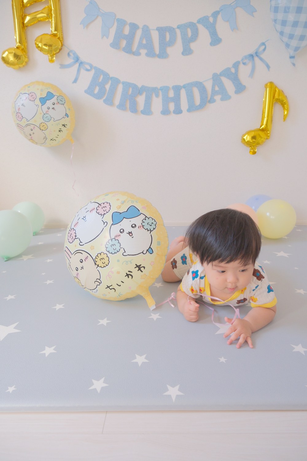 星柄のマットに手をついたお子さまが、床に置いたちいかわのバルーンのすぐそばに顔を寄せている。後ろに水色のHAPPY BIRTHDAYガーランドと金の音符