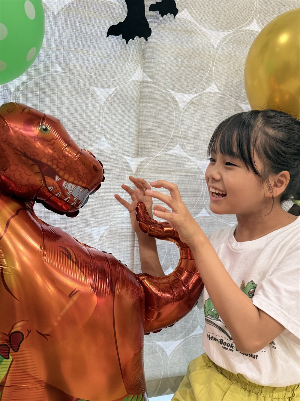 ティラノサウルスのバルーンと向かい合って手を伸ばしているお子さま。背景はレースのカーテンで、顔にも飾りにも強い影が出ていない