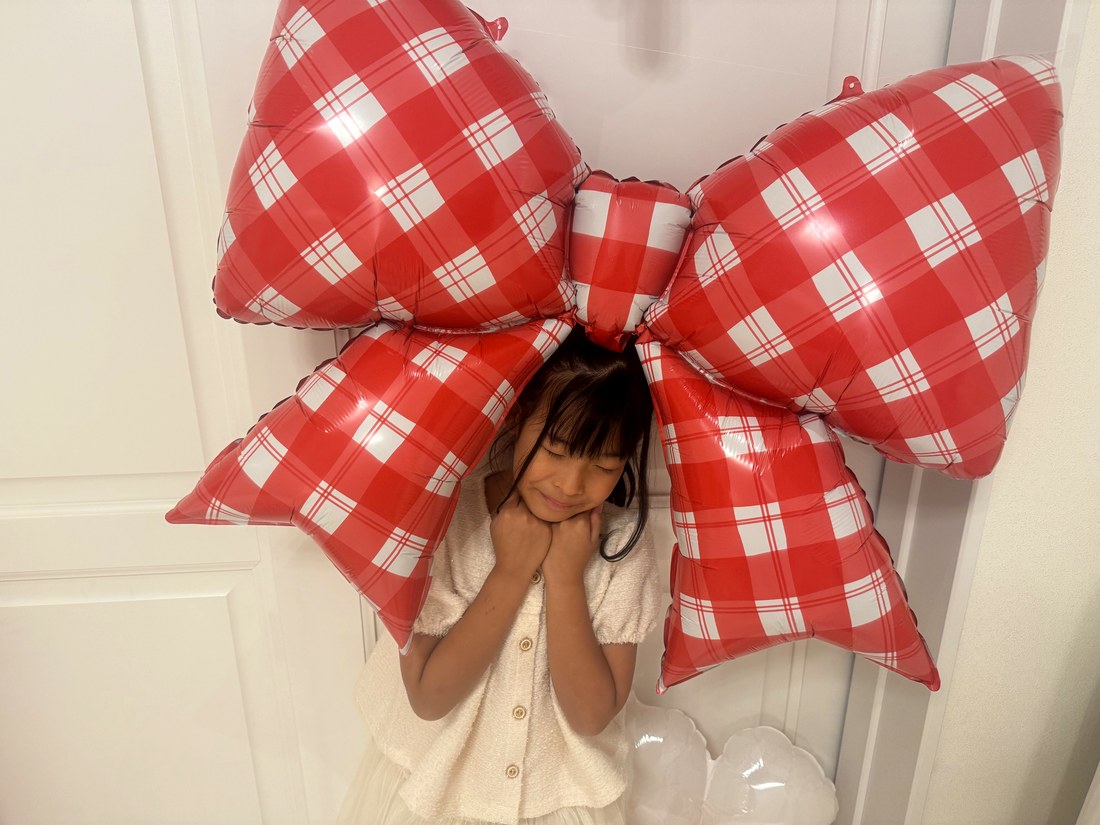 赤いギンガムチェックの大きなリボンのバルーンの真下に、クリーム色のお洋服のお子さまが立ち、両手をほおに当てて目を閉じている。リボンが頭の上にのっているように見える