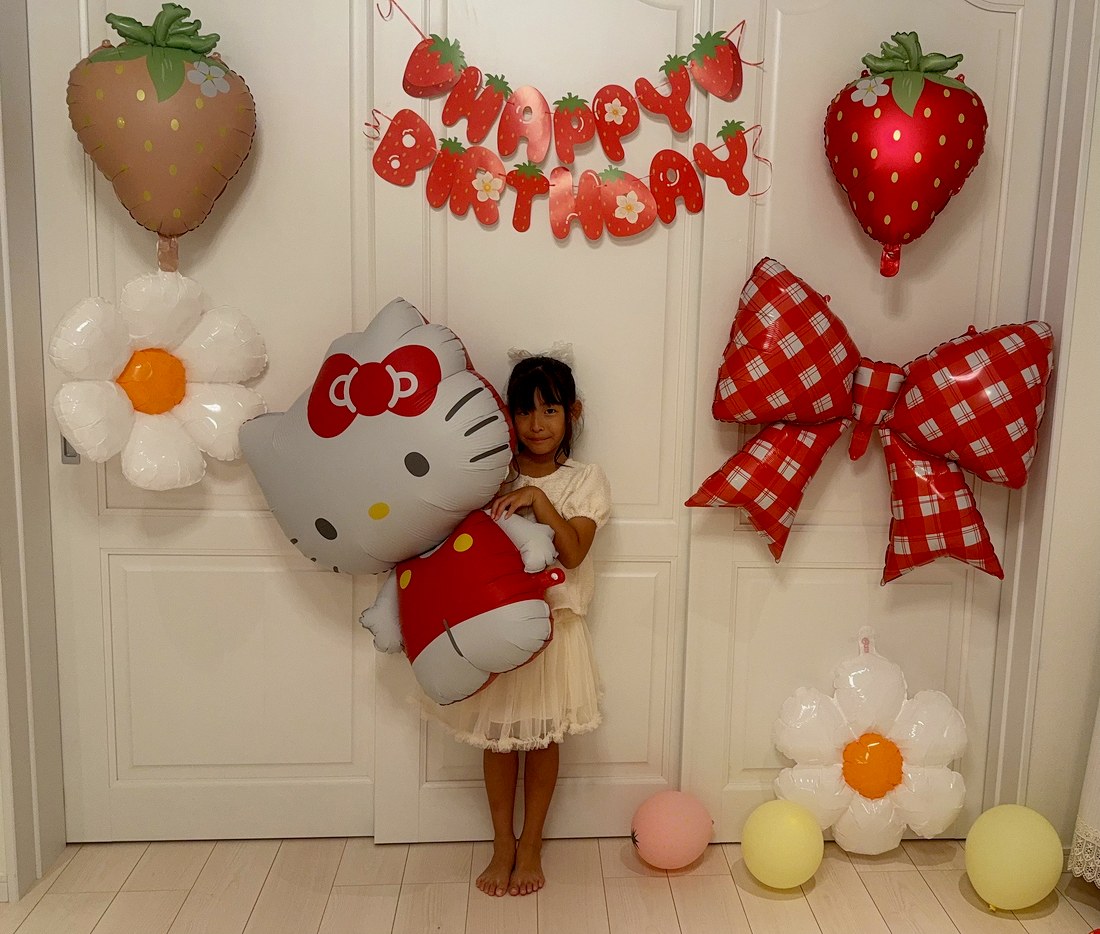 いちごキティの飾り付けの前で、クリーム色のブラウスとチュールスカートのお子さまが、ハローキティのバルーンを抱えてほほえんでいる。左上にピンクのいちご、右に赤いチェックのリボン