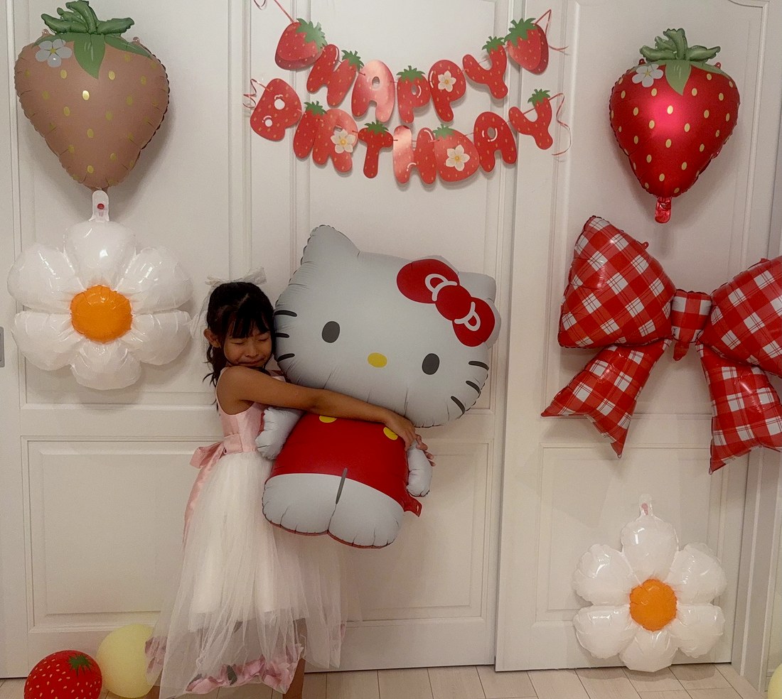 ピンクと白のチュールのドレスを着たお子さまが、ハローキティのバルーンを両手で抱きしめ、目を閉じてほほえんでいる。まわりにデイジー、いちご、赤いチェックのリボン