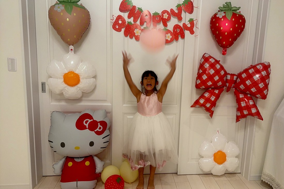 いちごキティの飾り付けの前で、ピンクと白のドレスのお子さまが両手を上げ、ピンクの風船を投げ上げている。左にハローキティ、右に赤いチェックのリボンとデイジー