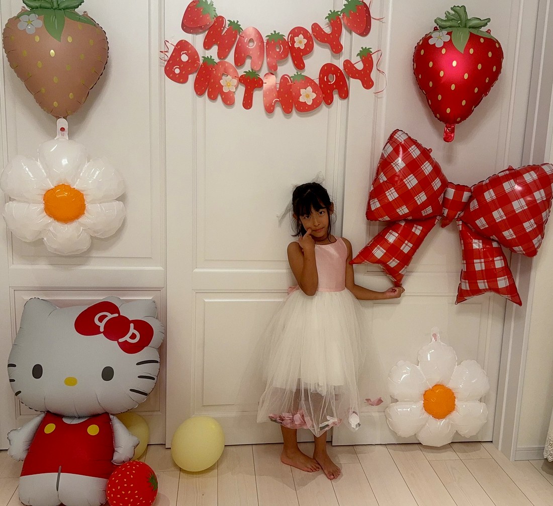 いちごキティの飾り付けの全体。上にいちごのHAPPY BIRTHDAYガーランドとピンクと赤のいちご、左にデイジーと床に置いたハローキティ、右に赤いチェックのリボンとデイジー。真ん中にピンクと白のドレスのお子さまが立ち、ほおに指を当てている
