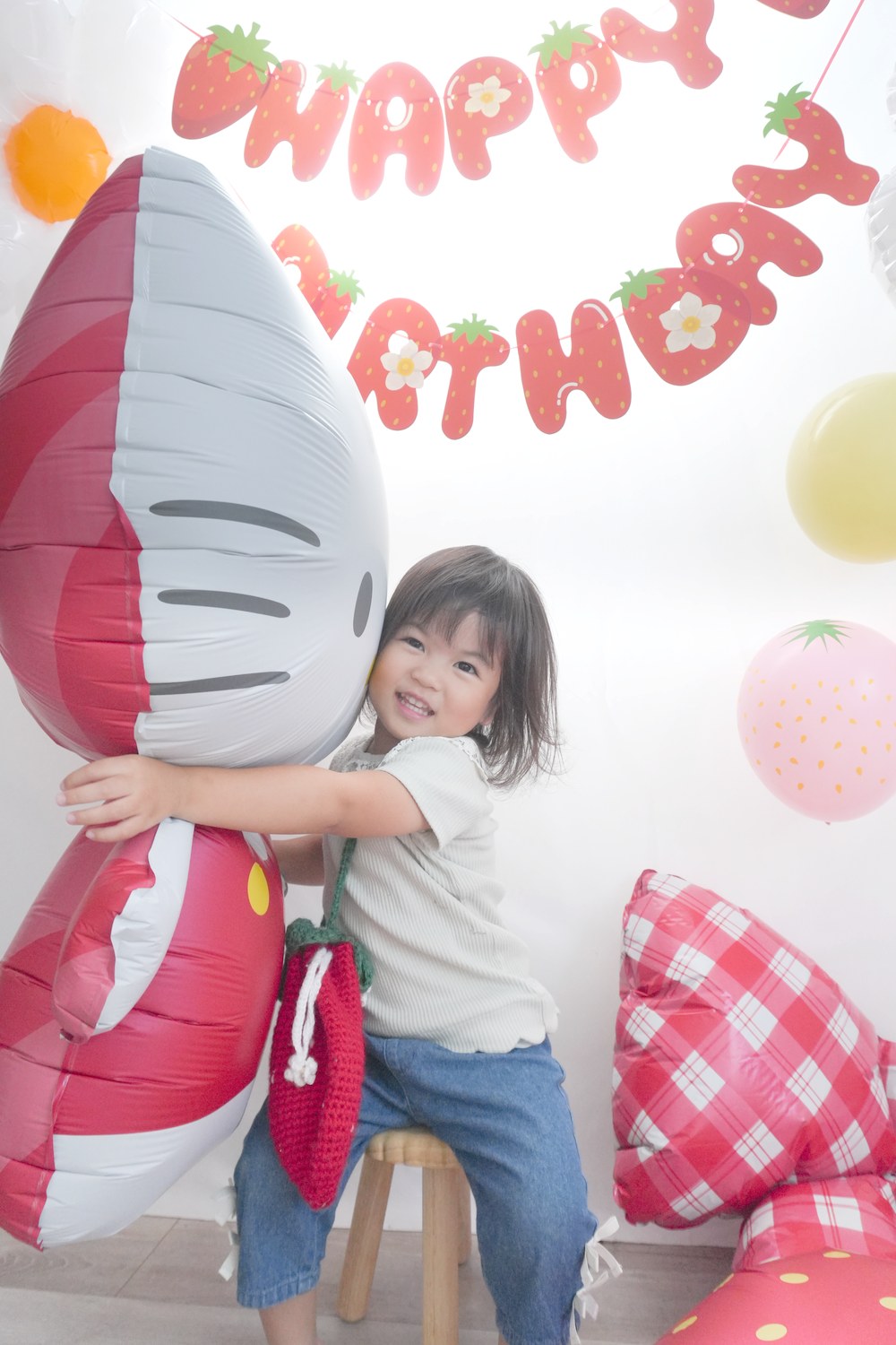 ハローキティのバルーンに横から抱きついて笑うお子さま。肩から赤い編みのポシェットを下げている。後ろにいちご柄のHAPPY BIRTHDAYガーランドと、赤いギンガムチェックのリボン