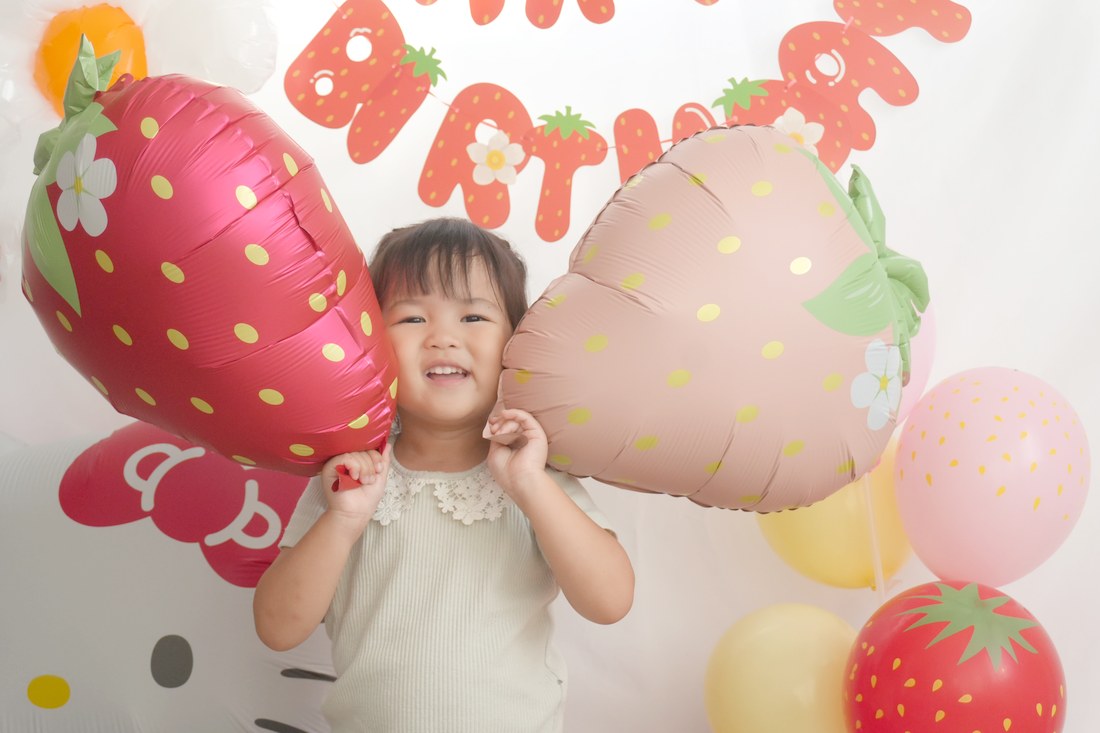 赤いいちごとピンクのいちごのバルーンのあいだに顔を入れて、両手をほっぺに当てて笑うお子さま。後ろにいちご柄のガーランド