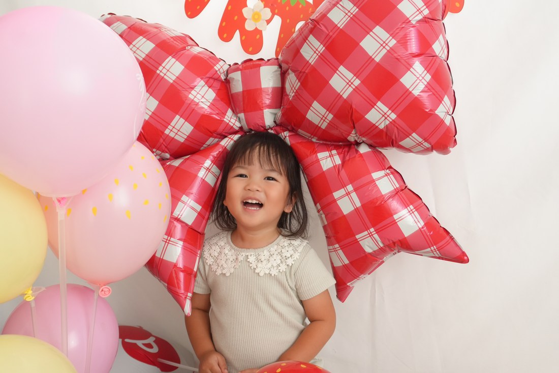赤いギンガムチェックの大きなリボンのバルーンの前で、お子さまが口を大きく開けて笑っている。左にピンクと黄色のゴム風船