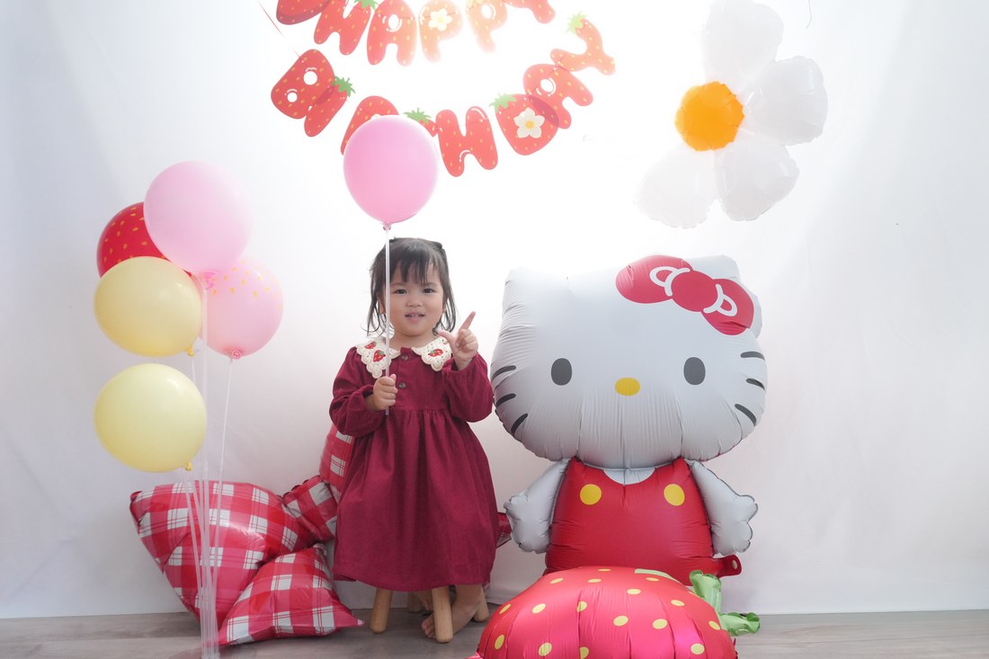 赤いワンピースに着替えたお子さまが、ハローキティのバルーンの隣に立って、ピンクの風船を手に持っている。上にいちご柄のHAPPY BIRTHDAYガーランドとデイジー、足もとにいちごとチェックのリボン