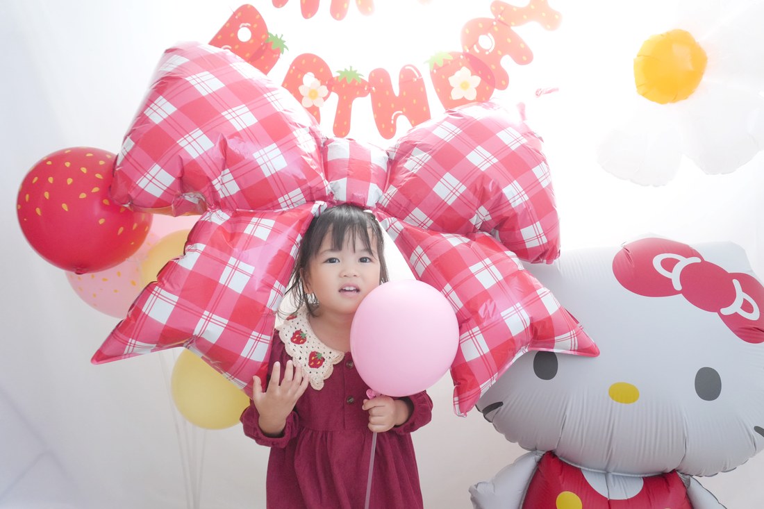 赤いワンピースのお子さまが、赤いギンガムチェックのリボンの前でピンクの風船を持っている。右にハローキティのバルーン、左に赤いいちご柄のゴム風船