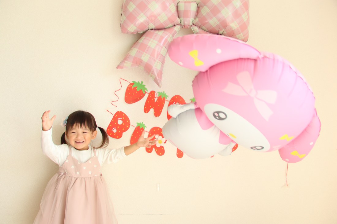マイメロディのバルーンが頭の上にきて、お子さまが両手を上げて笑っている。背景にチェックのリボンといちご柄のガーランド