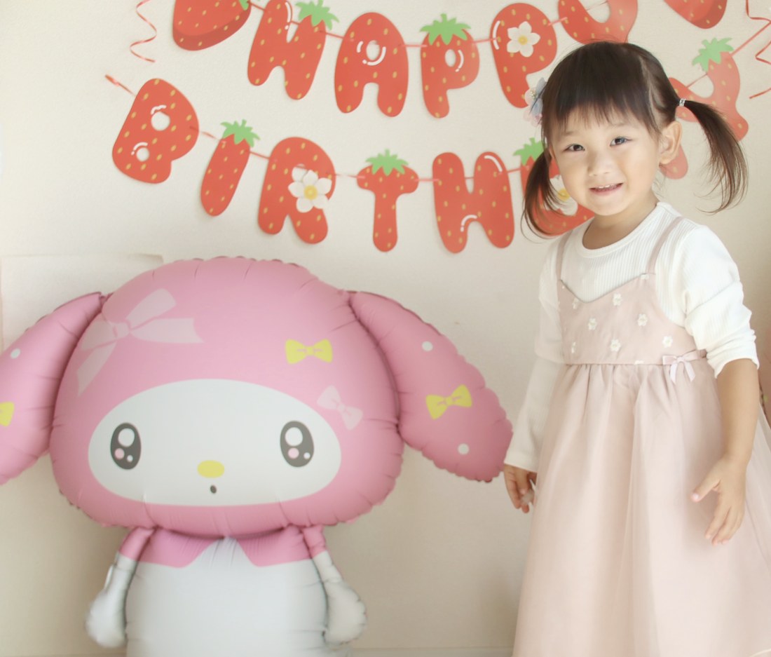 床に置いたマイメロディのバルーンの右隣に、ピンクのチュールのお洋服のお子さまが立って笑っている。背景にはいちご柄のHAPPY BIRTHDAYガーランド
