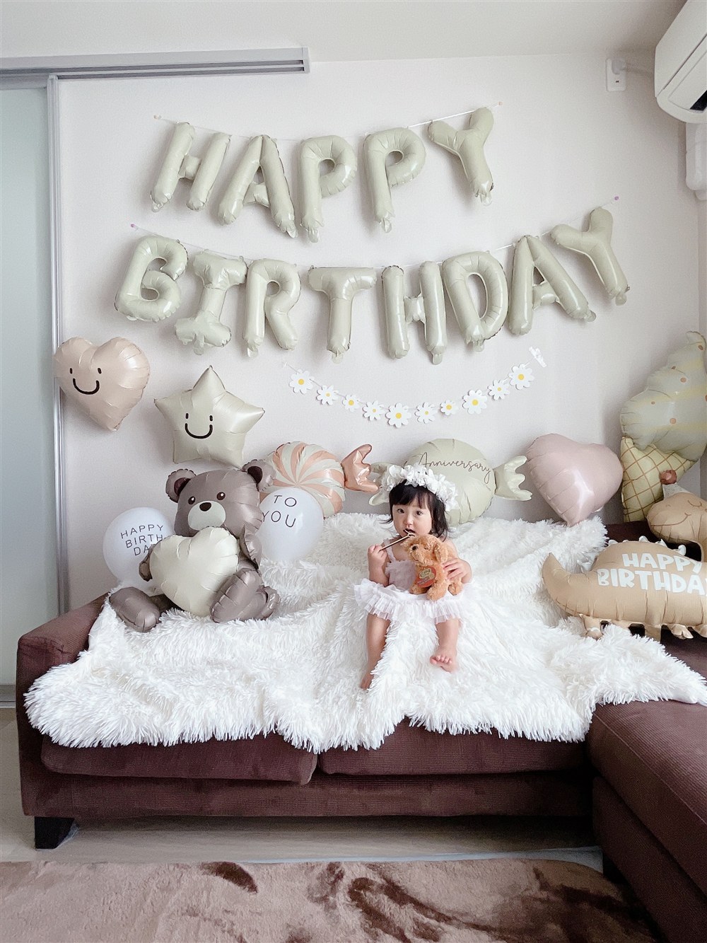 白い壁とブラウンのソファのリビングで、くすみカラーの飾り付けをしたところ。ベージュのHAPPY BIRTHDAYレターバルーン、ブラウンのくまのバルーン、ベージュのハートと星のバルーン、白いお花のガーランドが壁とソファの上に並んでいる