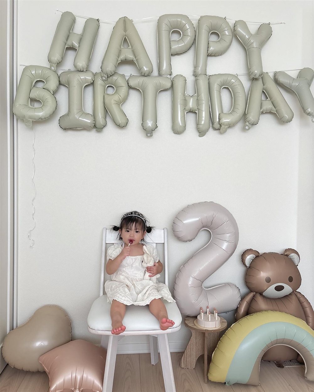 白い壁にくすみグリーンのHAPPY BIRTHDAYの文字バルーン、その下に数字2のバルーン、くまのバルーン、虹のバルーン、ハートと星のバルーン。白い椅子に座った子どもが飴を持っている