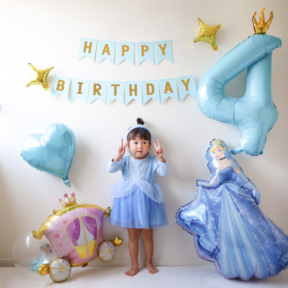 水色のHAPPY BIRTHDAYガーランドの下で、水色のドレスを着た子どもがピースをしている。横に大きな数字4のバルーンとシンデレラのバルーン、かぼちゃの馬車のバルーン