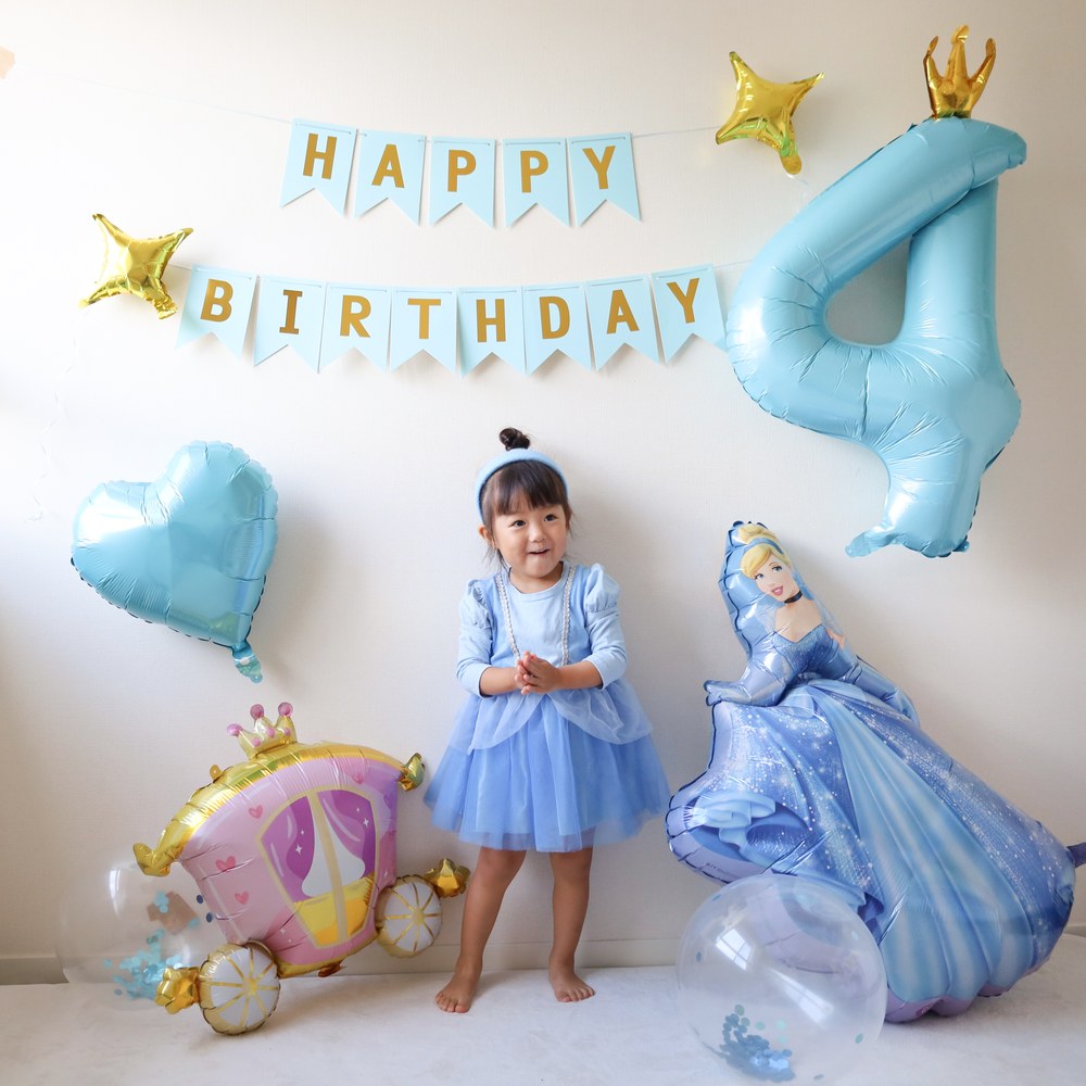 水色のHAPPY BIRTHDAYガーランドの下で、水色のドレスを着た子どもが手を合わせて笑っている。横に大きな数字4のバルーンとシンデレラのバルーン、かぼちゃの馬車のバルーン