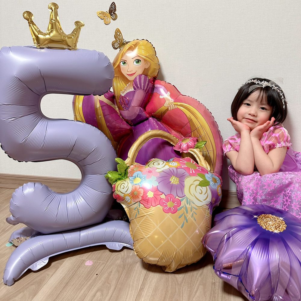蝶のHAPPY BIRTHDAYガーランドの下で、紫のドレスの子どもがほおづえをついて笑っている。横に王冠をのせた紫の数字5のバルーンと、ラプンツェルのバルーン、花かごのバルーン