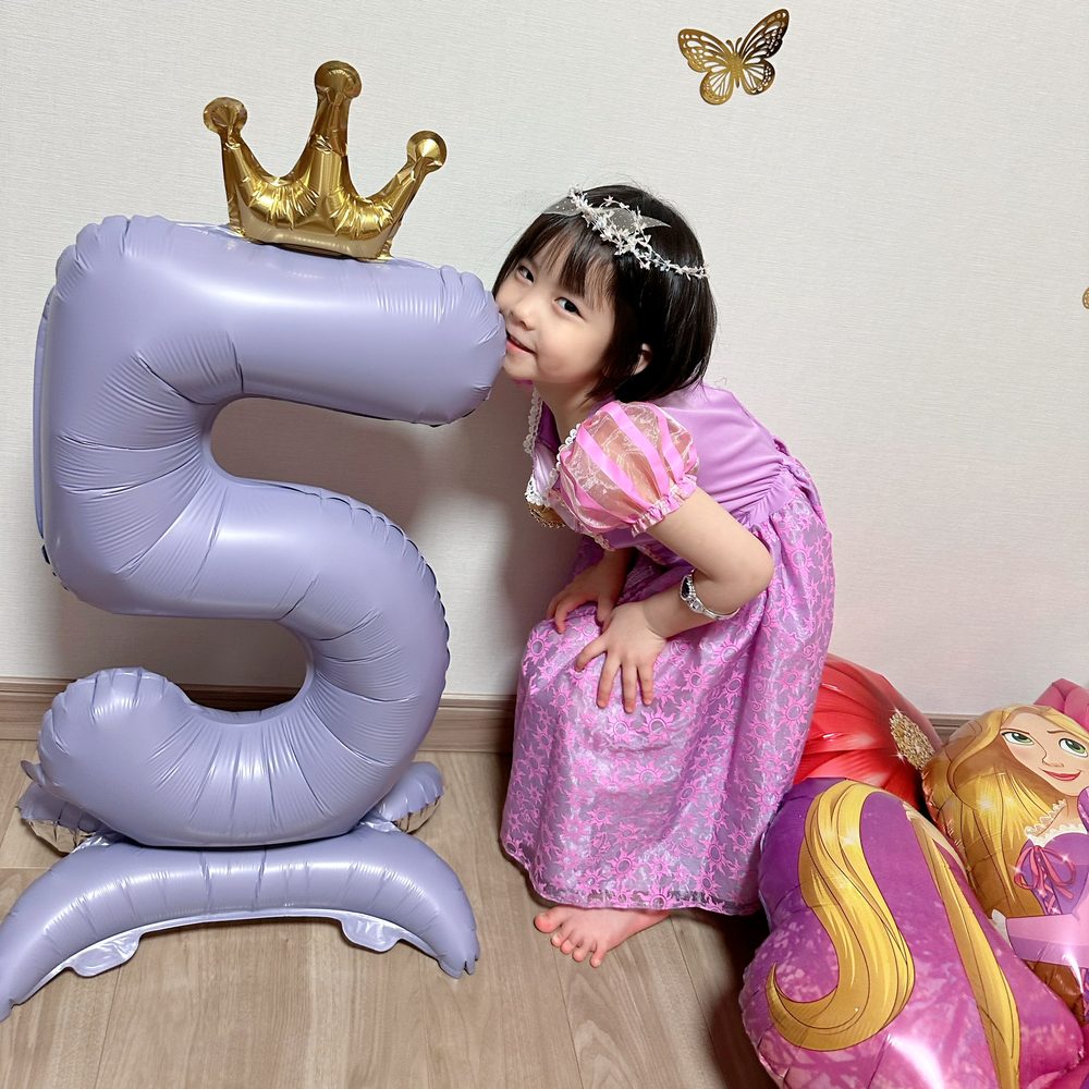 王冠をのせた紫の数字5のバルーンに、ドレスの子どもが顔を寄せて笑っている。足元にラプンツェルのバルーン