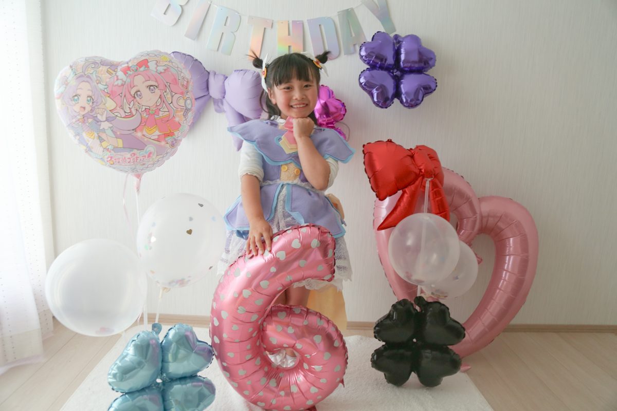 パステルカラーのHAPPY BIRTHDAYガーランドの下で、子どもがピンクに白いハート柄の数字6のバルーンに手をのせて笑っている。横にプリキュアのハート型バルーン、紫と黒のクローバー、赤いリボンのバルーン