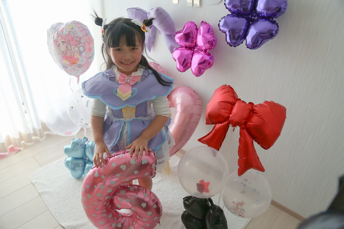 ピンクに白いハート柄の数字6のバルーンに手をのせて笑う子ども。うしろにプリキュアのハート型バルーン、赤いリボンのバルーン、紫とピンクのクローバー