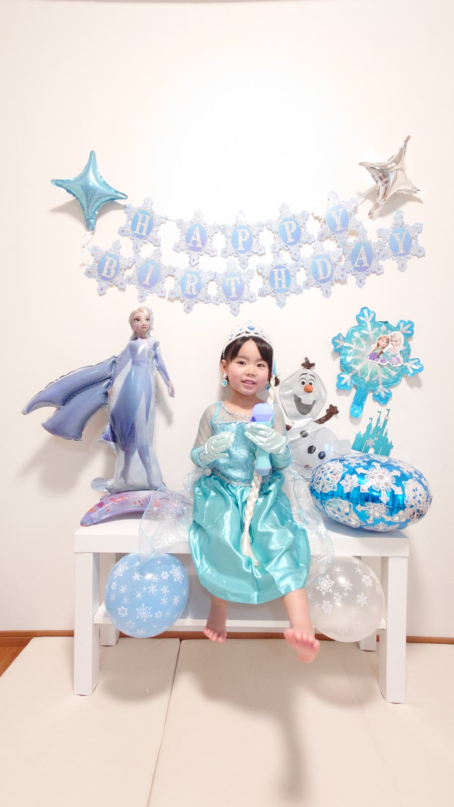 エルサのドレスとティアラを身につけた女の子と、自立式のエルサのバルーン、オラフのバルーン、雪の結晶のHAPPY BIRTHDAYガーランド、雪の結晶柄のゴム風船の誕生日飾り付け