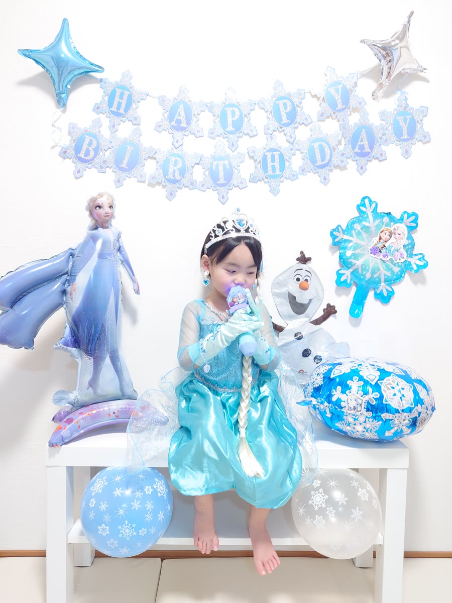 エルサのドレスを着てティアラをつけた女の子が、オラフと雪の結晶のバルーンに囲まれて座っている誕生日の飾り付け実例