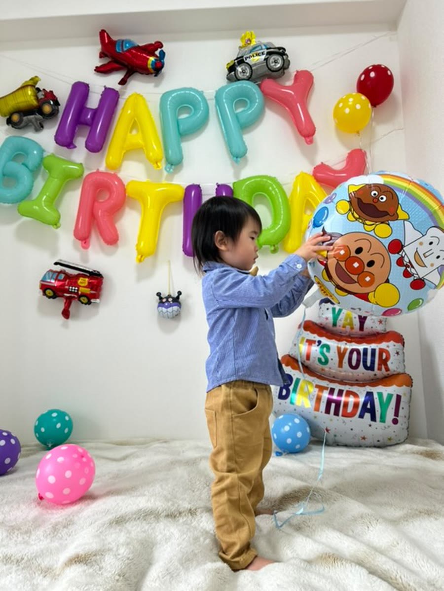 アンパンマンの丸いバルーンに手を伸ばす小さな子。カラフルなHAPPY BIRTHDAYの文字バルーンと飛行機・パトカー・消防車の乗り物バルーン、水玉のゴム風船