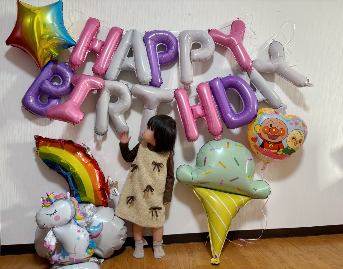 パステルカラーのHAPPY BIRTHDAY文字バルーンとユニコーン・虹・アイスクリームのバルーン、アンパンマンのハート型バルーンを見上げる小さな女の子