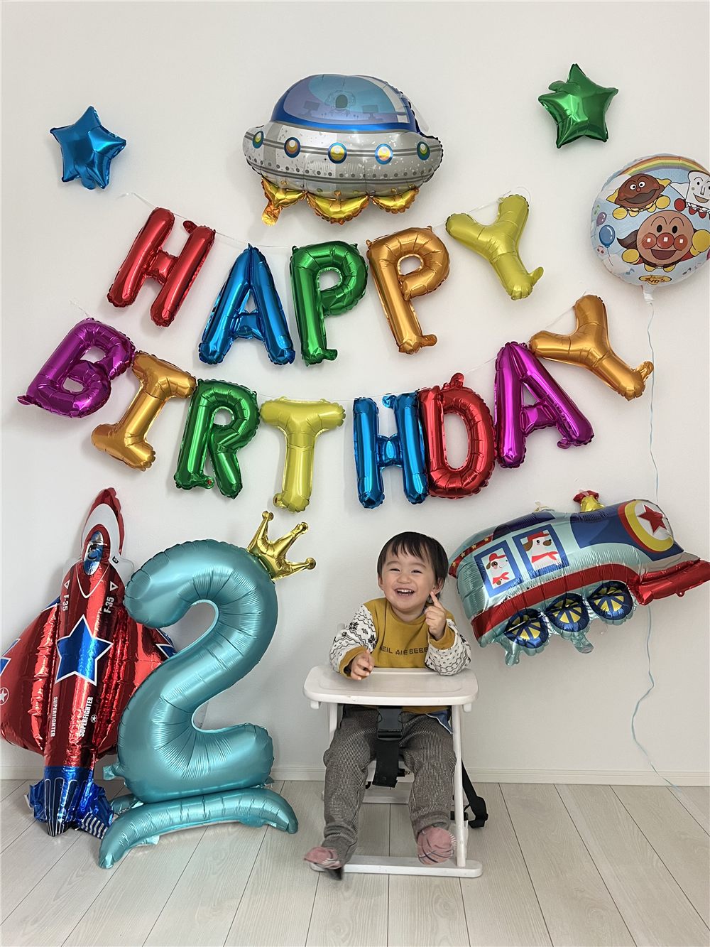 白い壁にHAPPY BIRTHDAYの文字バルーンが2段、その上にUFO、右に丸いアンパンマンのバルーンと機関車、左の床に赤い飛行機と王冠つきの数字2のバルーン。ベビーチェアに座った子どもが笑っている