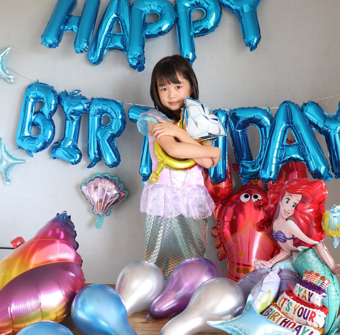 青いHAPPY BIRTHDAYのバルーンの前で、人魚のドレスを着た子どもがバルーンを抱えて立っている。右にアリエルのバルーン、手前に海のモチーフのバルーンとメタリックのゴム風船