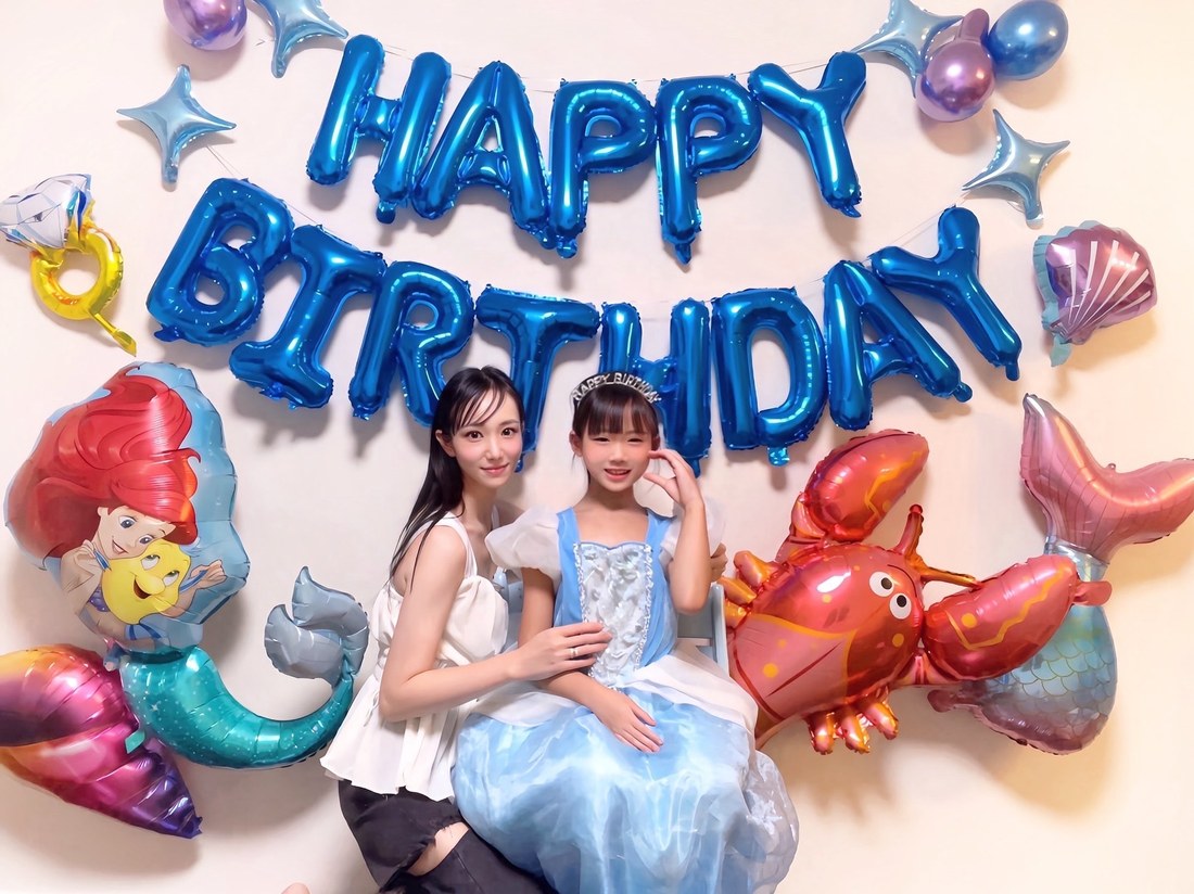 青いHAPPY BIRTHDAYのバルーンの下、アリエルのバルーンと海のモチーフのバルーンに囲まれて、水色のドレスの子どもと大人が並んで座っている