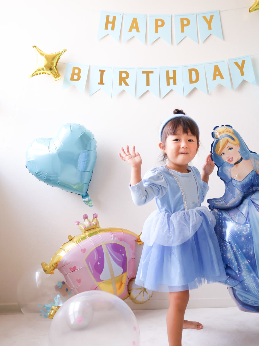 シンデレラのドレスを着てシンデレラのバルーンの隣で手を振る3歳くらいの女の子。水色のハートと馬車のバルーン、HAPPY BIRTHDAYガーランド