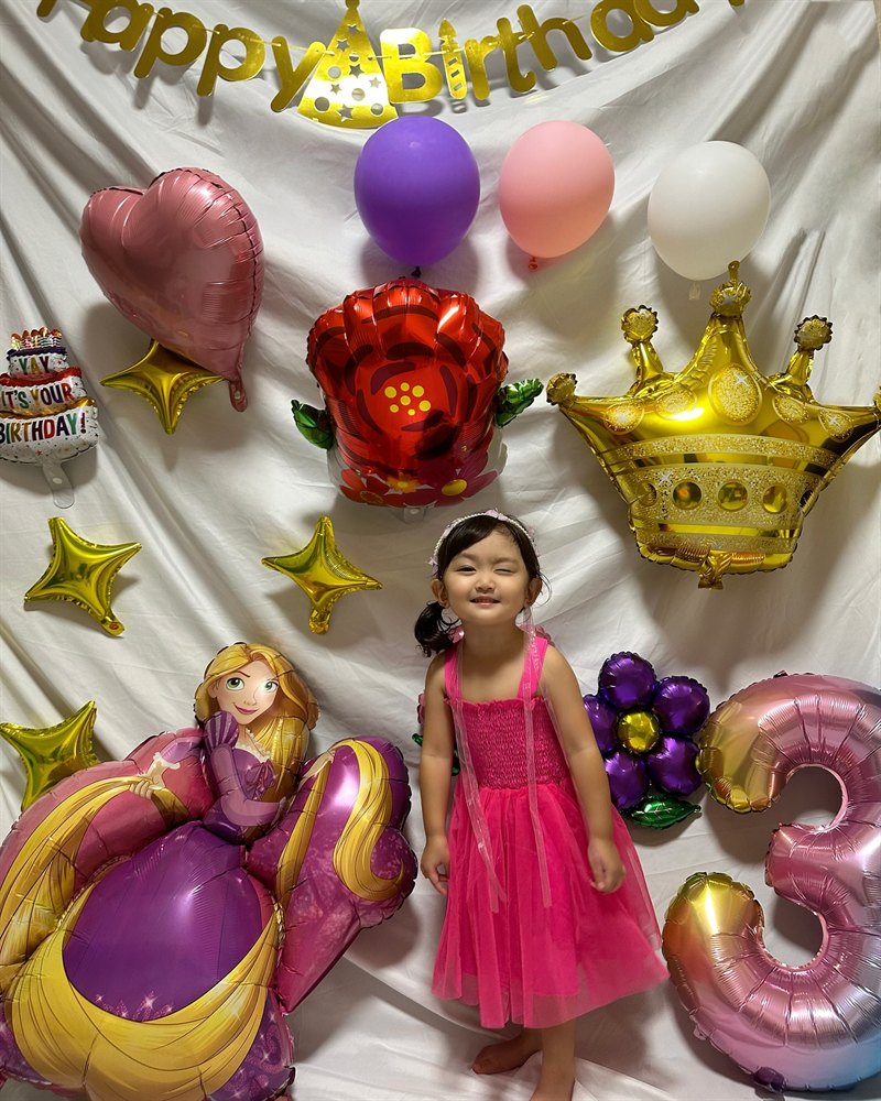 白い布を背にした誕生日の飾り付け。上のほうに金色の筆記体のHappy Birthdayガーランドが横に渡してあり、その下にラプンツェルのバルーンを中心に、赤い花・王冠・星のバルーンが並んでいる