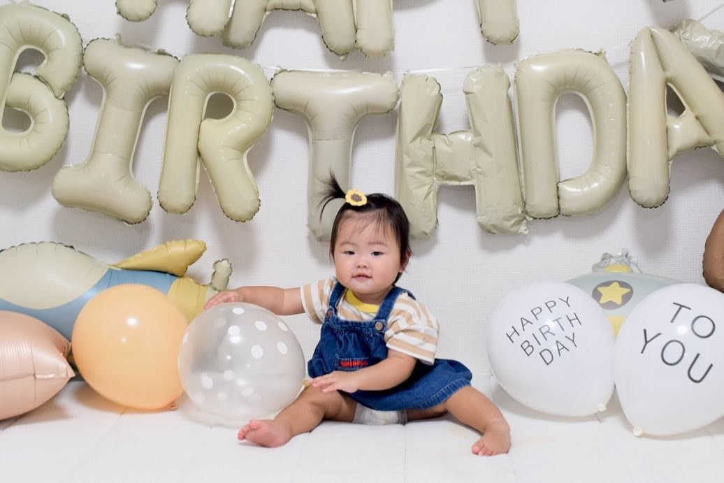くすみカラーのHAPPY BIRTHDAYの文字バルーンの下で、床に座った子どもが透明に白い水玉のゴム風船に手をのばしている。左に飛行機とオレンジのバルーン、右にHAPPY BIRTH DAYとTO YOUと書かれた白い風船