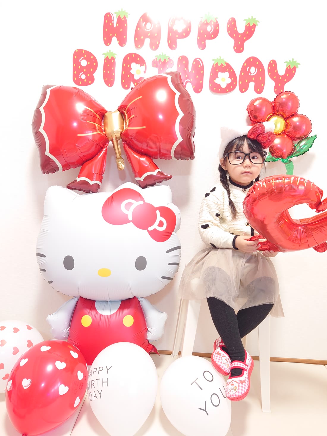 HAPPYとBIRTHDAYのいちごの紙ガーランドを1枚ずつ壁に貼って飾った実例。特大ハローキティバルーンと赤いリボンバルーン、数字の6のバルーンを持って座る女の子