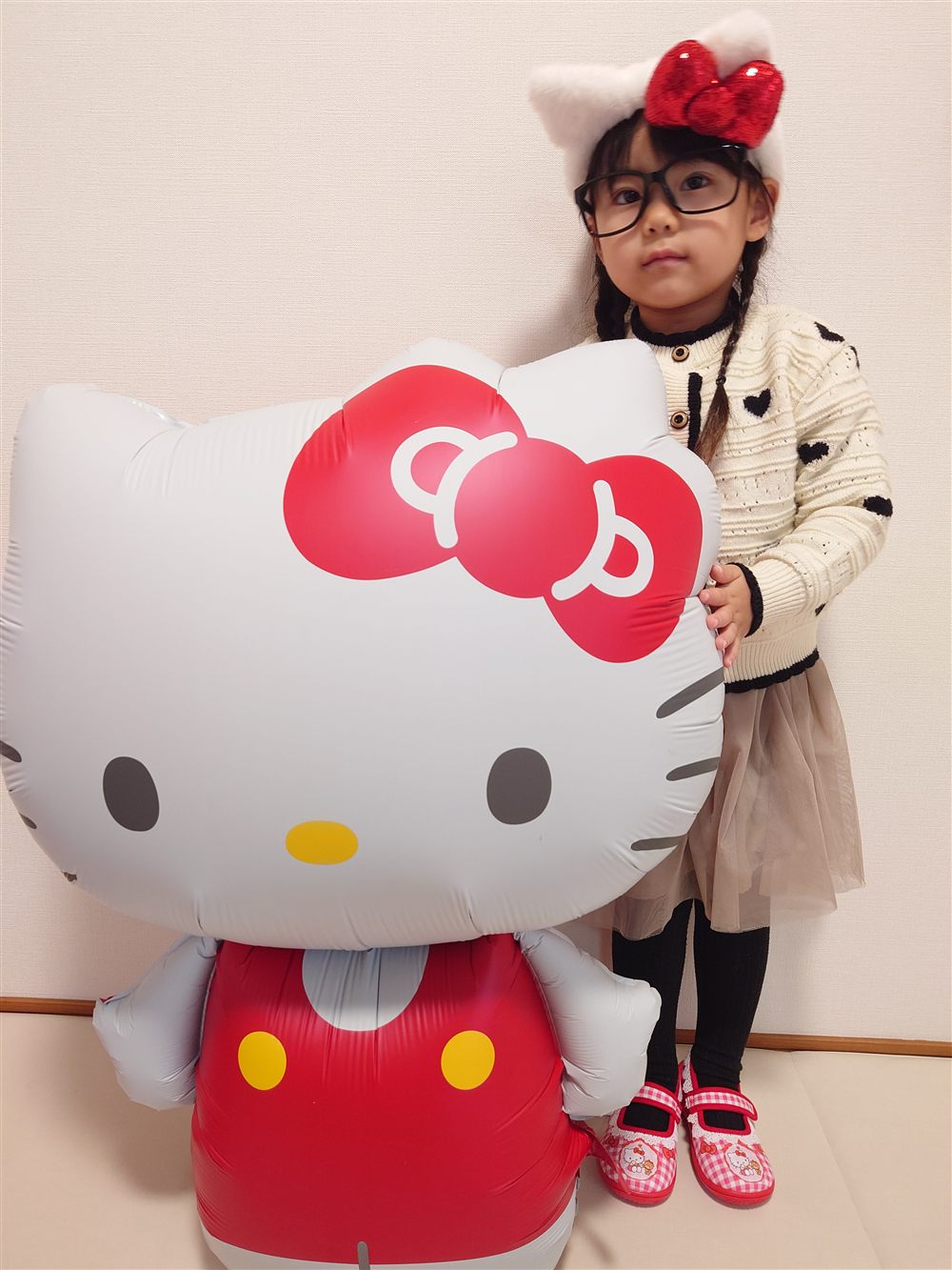 キティの耳と赤いリボンのカチューシャをつけた子どもが、特大ハローキティバルーンの横に立っている