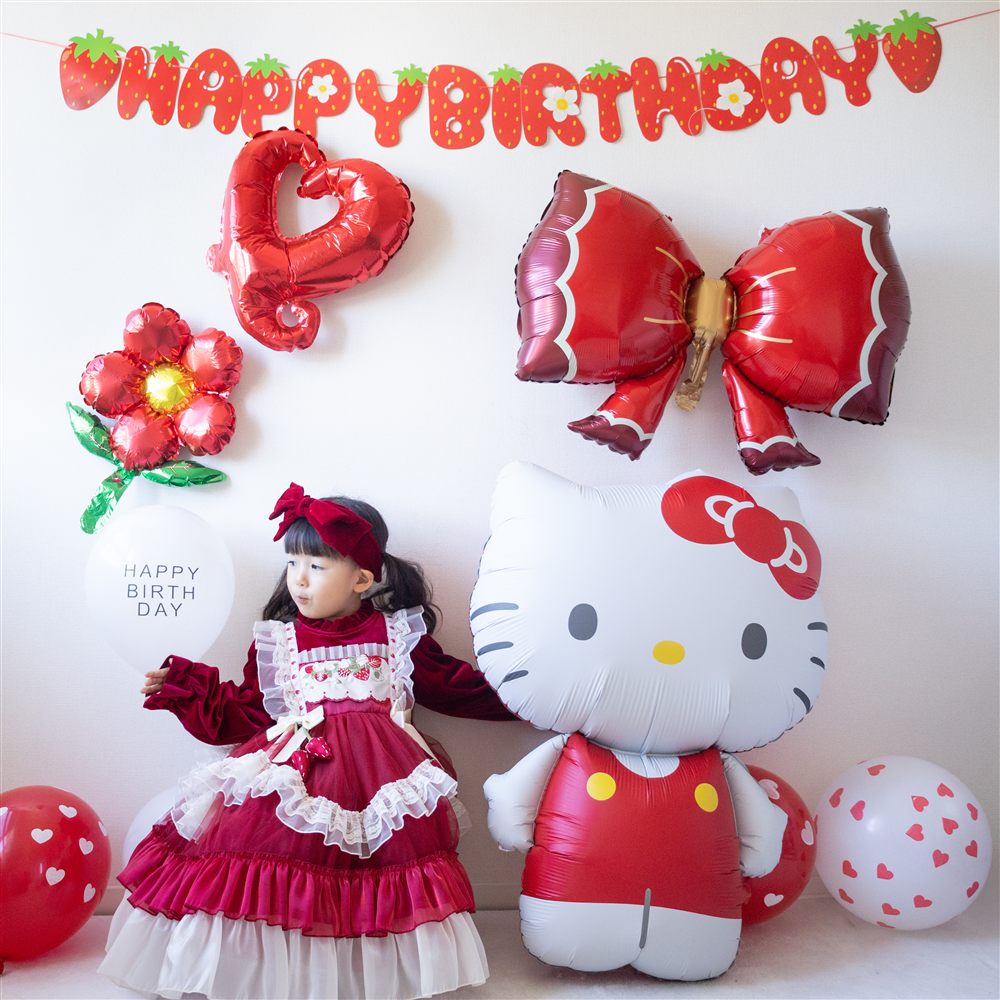 いちごのHAPPY BIRTHDAYガーランドの下に、赤いハート・リボン・お花のバルーン。赤いワンピースの子どもが白いゴム風船を持ち、特大ハローキティバルーンの横に立っている