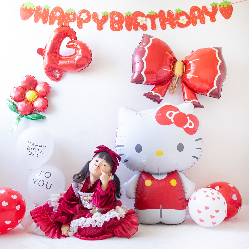 いちご柄のHAPPY BIRTHDAYガーランドの下に、赤いリボンとハート、お花のバルーンと大きなハローキティのバルーン。赤いワンピースの子どもが横に座っている