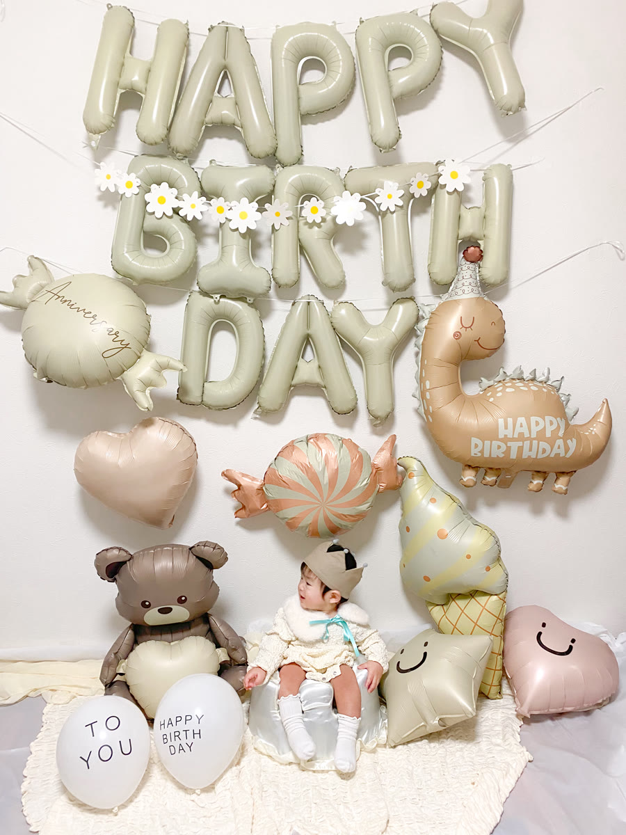 くすみカラーのHAPPY BIRTHDAYの文字バルーンを壁いっぱいに飾り、その下に「HAPPY BIRTH DAY」「TO YOU」と書かれた白いゴム風船を並べ、中央に帽子をかぶった小さな子が座っている誕生日の飾り付け
