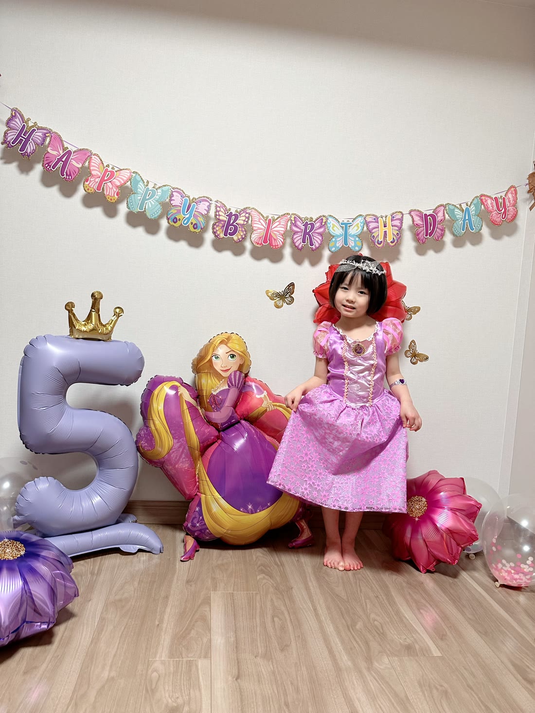 蝶々のHAPPY BIRTHDAYガーランドとラプンツェルバルーン、王冠付きのパープルの数字バルーン5の前に立つ、ラプンツェルのドレスの女の子