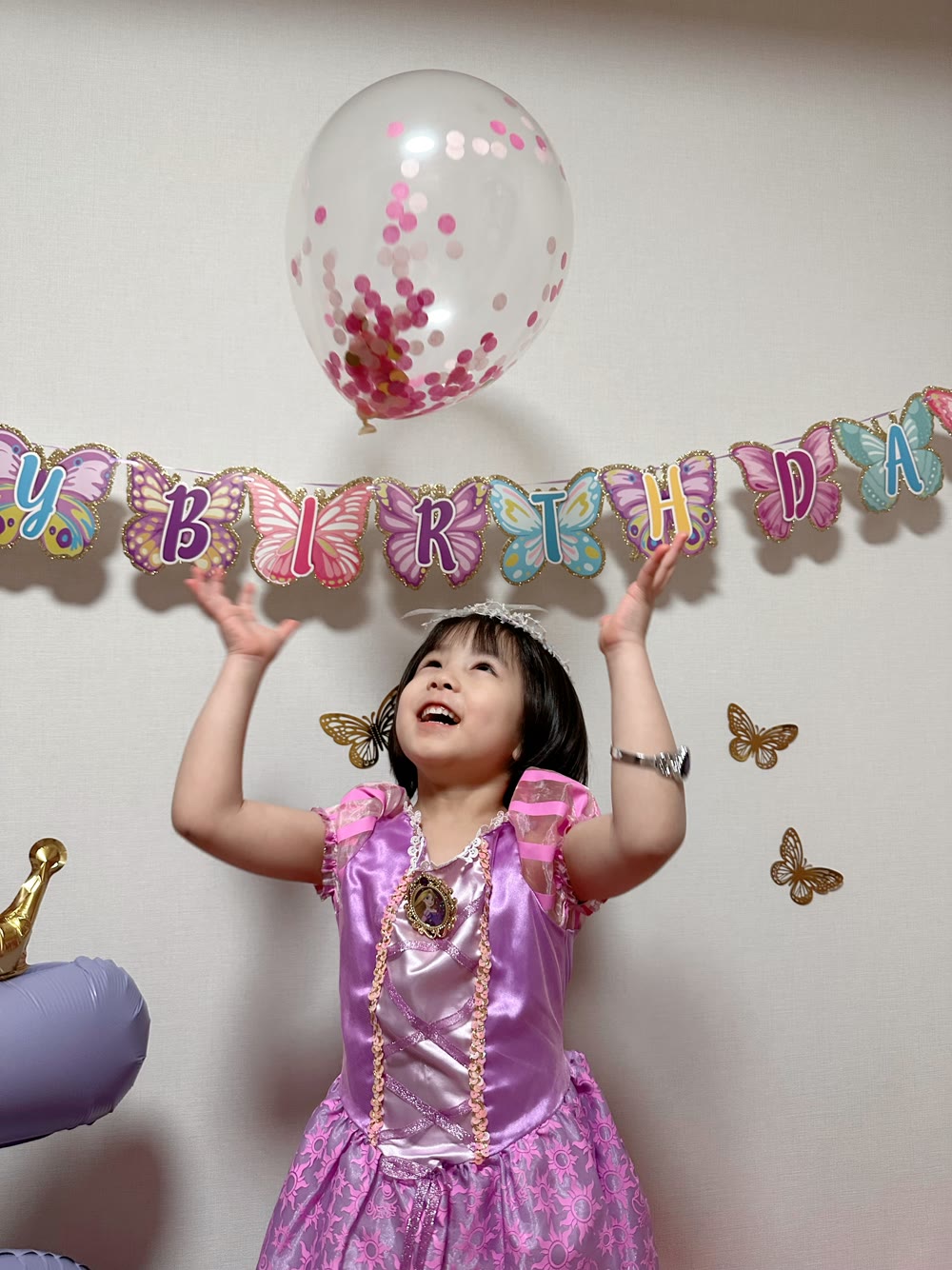金のラメで縁取られた蝶々のHAPPY BIRTHDAYガーランドの下で、コンフェッティバルーンを見上げて笑うラプンツェルのドレスの女の子