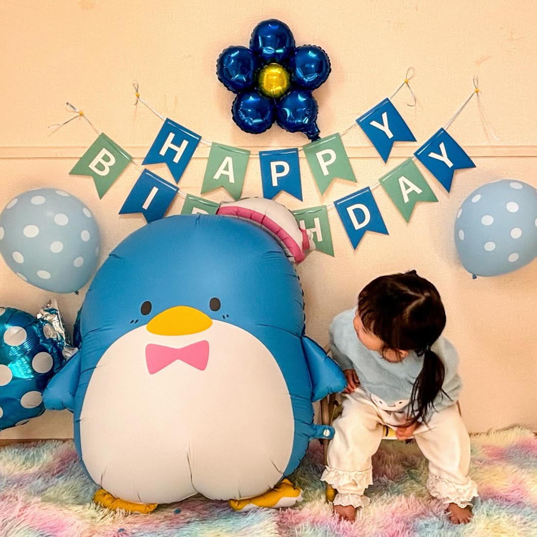 特大タキシードサムバルーンをのぞきこむ水色の服の女の子。ブルーのHAPPY BIRTHDAYガーランドとお花のバルーン、水玉のゴム風船