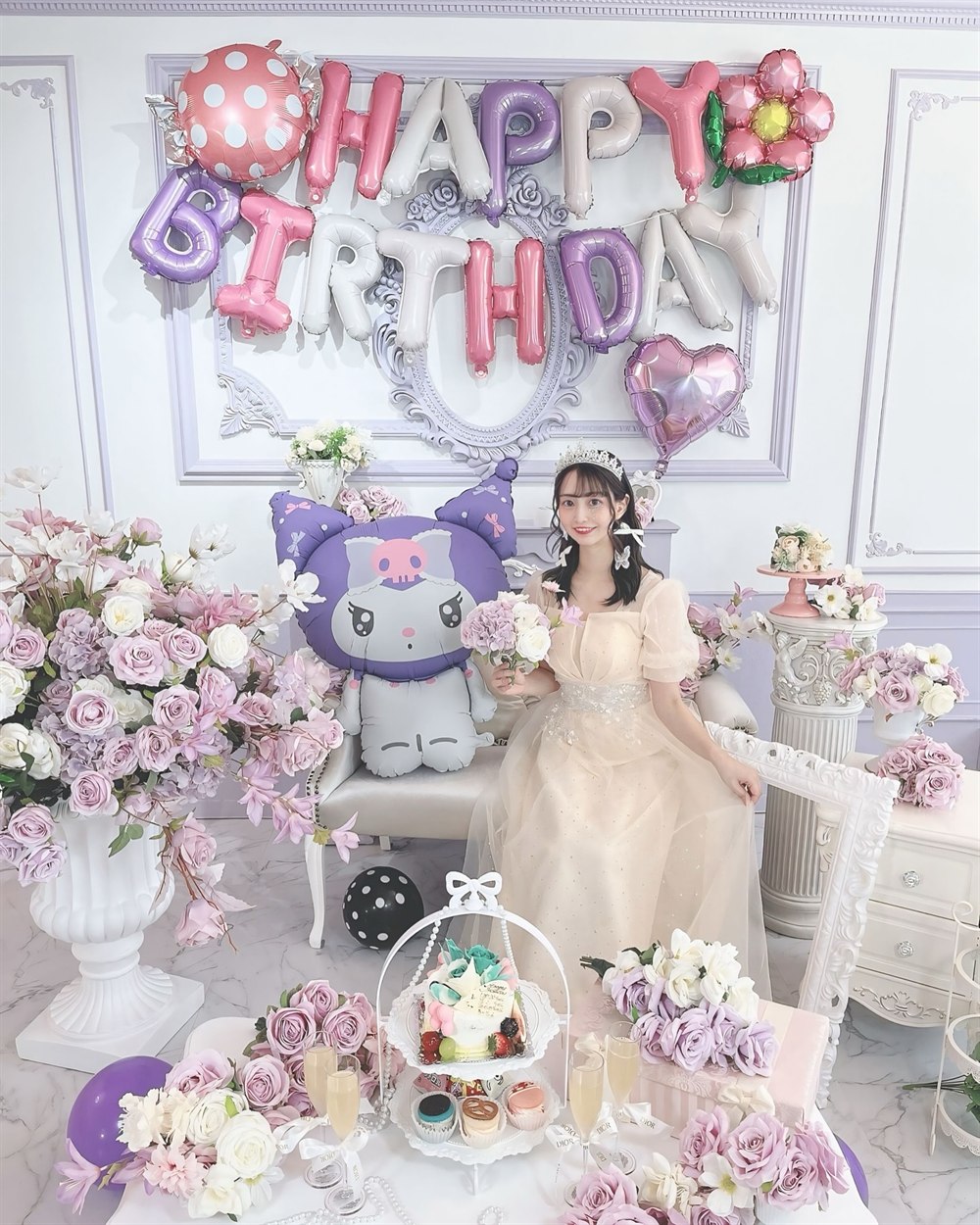 白とラベンダーの部屋で、ドレスを着た女性が特大クロミバルーンの隣に座り、HAPPY BIRTHDAYのレターバルーンと花に囲まれている大人の生誕祭の実例写真